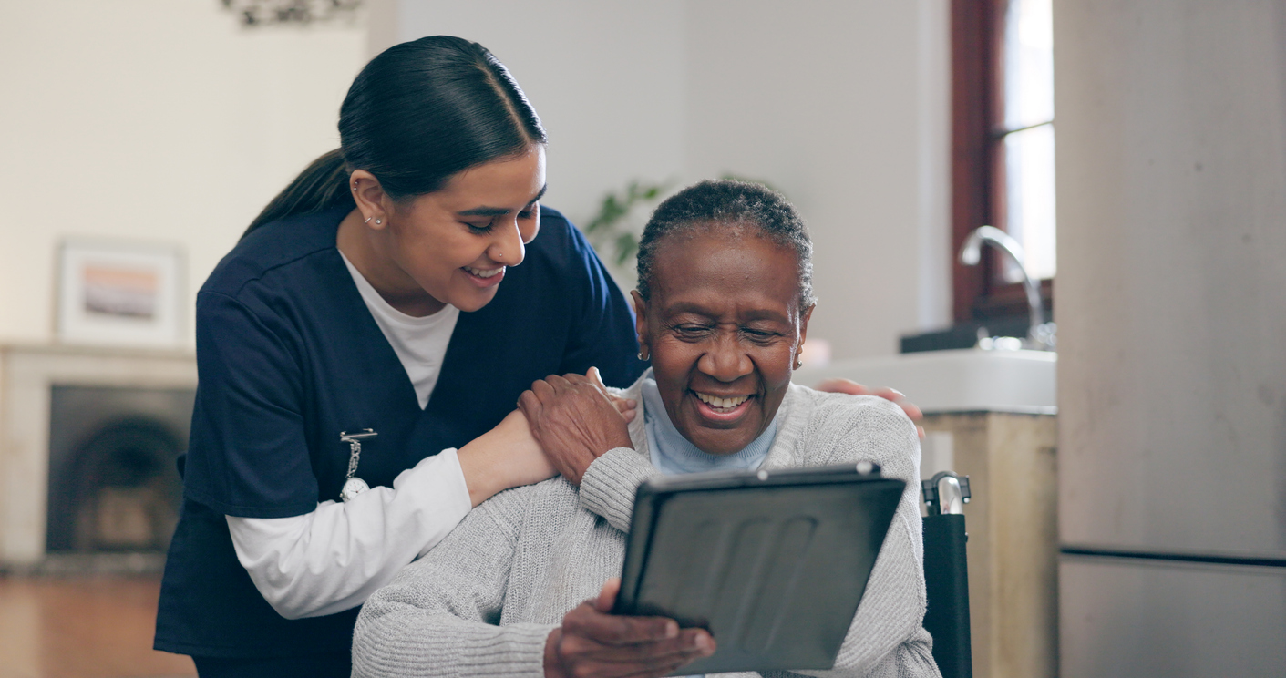 Female caregiver talks to senior black woman in rehabilitation clinic while both are looking at a tablet.