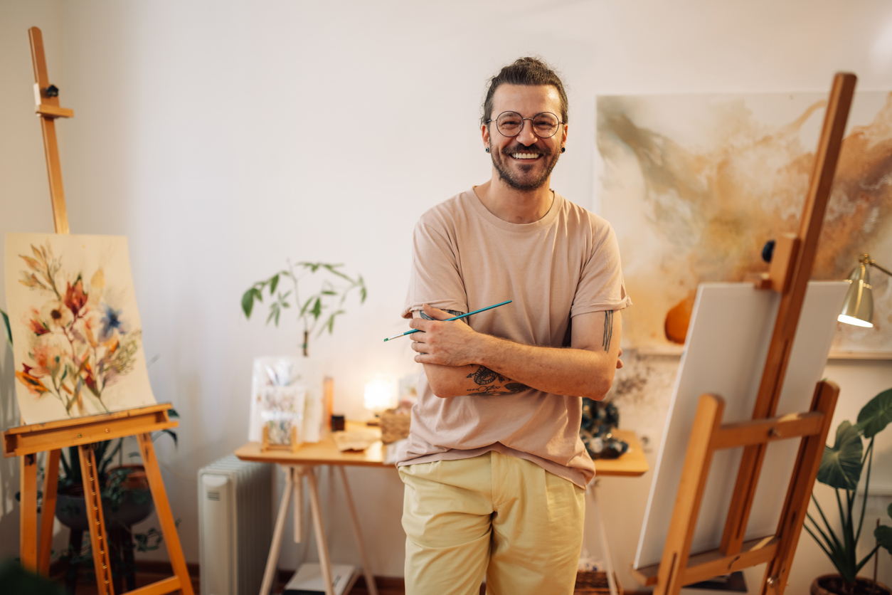 Portrait of a happy artistic tattooed trendy white male painter standing with arms crossed at creative atelier near easel and smiling at camera. Young creative hipster confident artist looking at camera at studio.