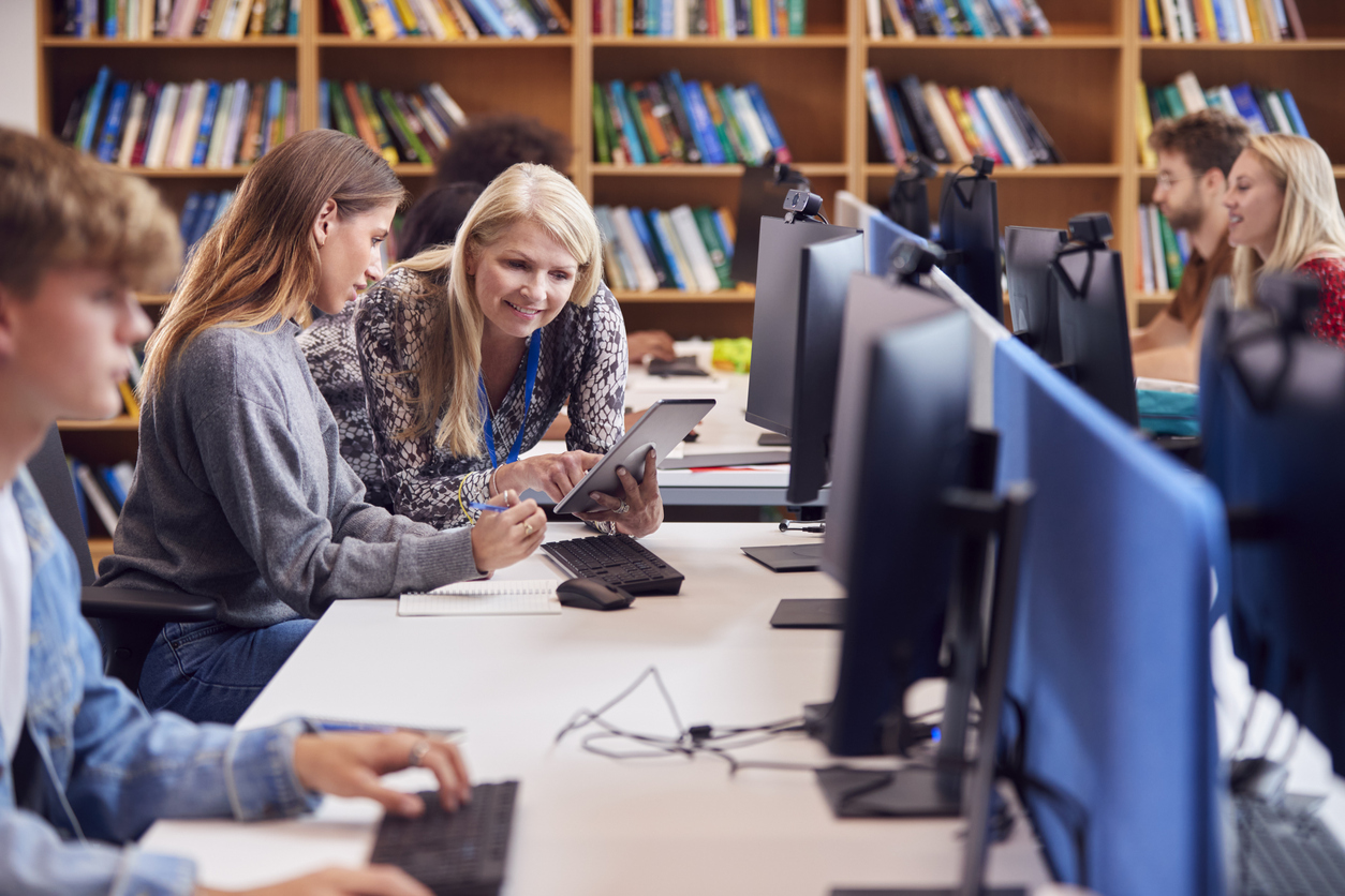 White female university or college student working at computer in library being helped by tutor who is using a tablet