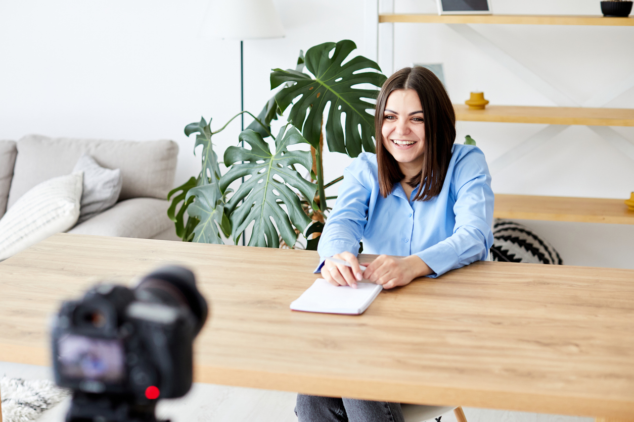 White female sitting at a desk being filmed with a corporate backdrop for a media company.
